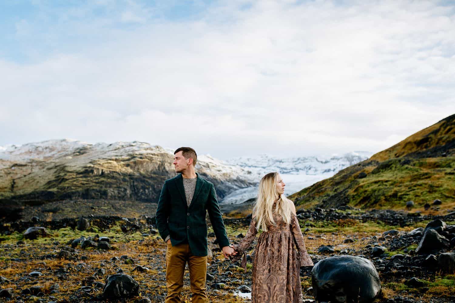 Man in brown pants and a jacket holds hands with a woman in a brown lace dress. They are looking in different directions.