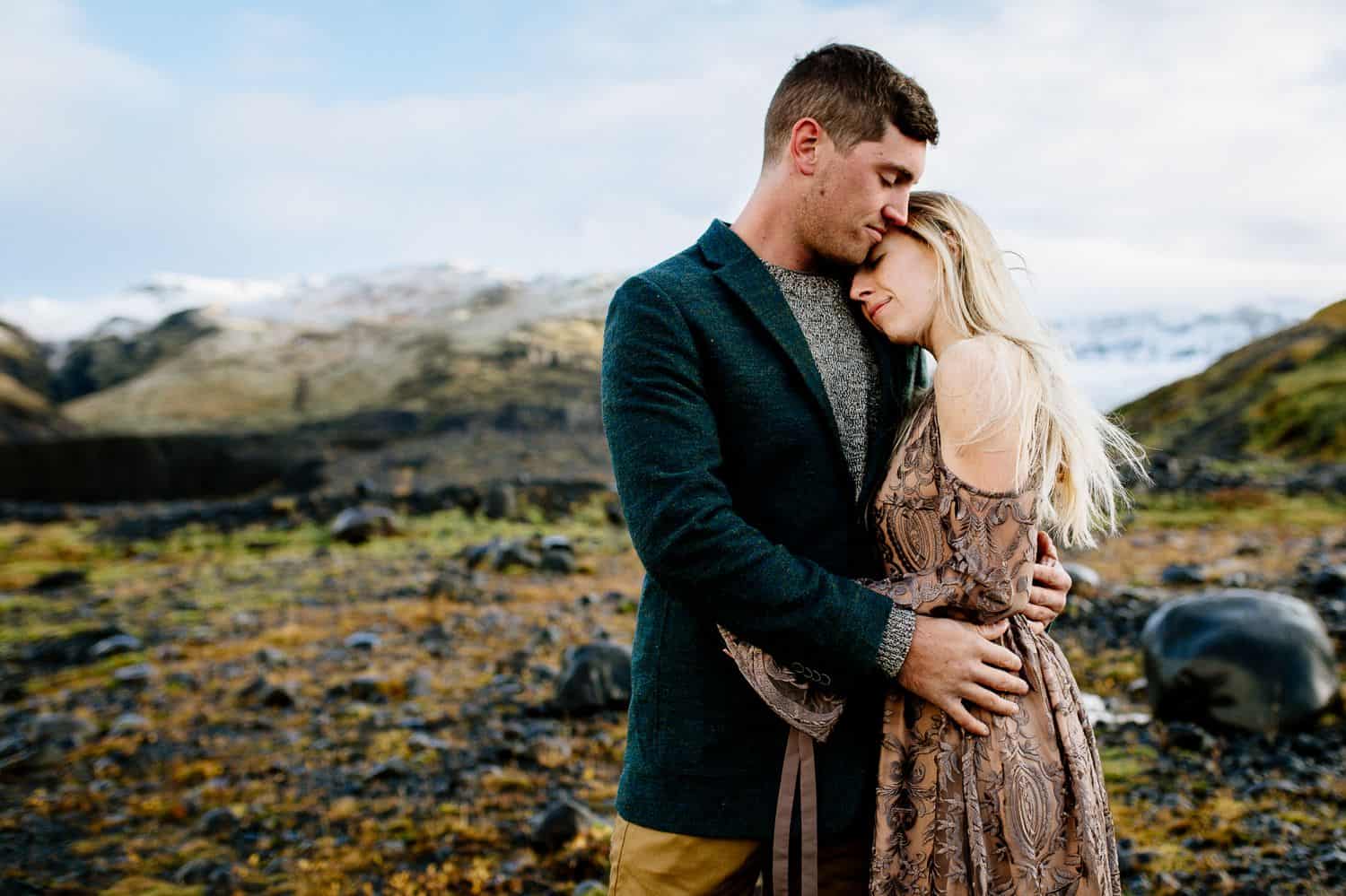 Man in a green jacket embraced a woman in a brown lace dress as they stand in the Icelandic landscape.