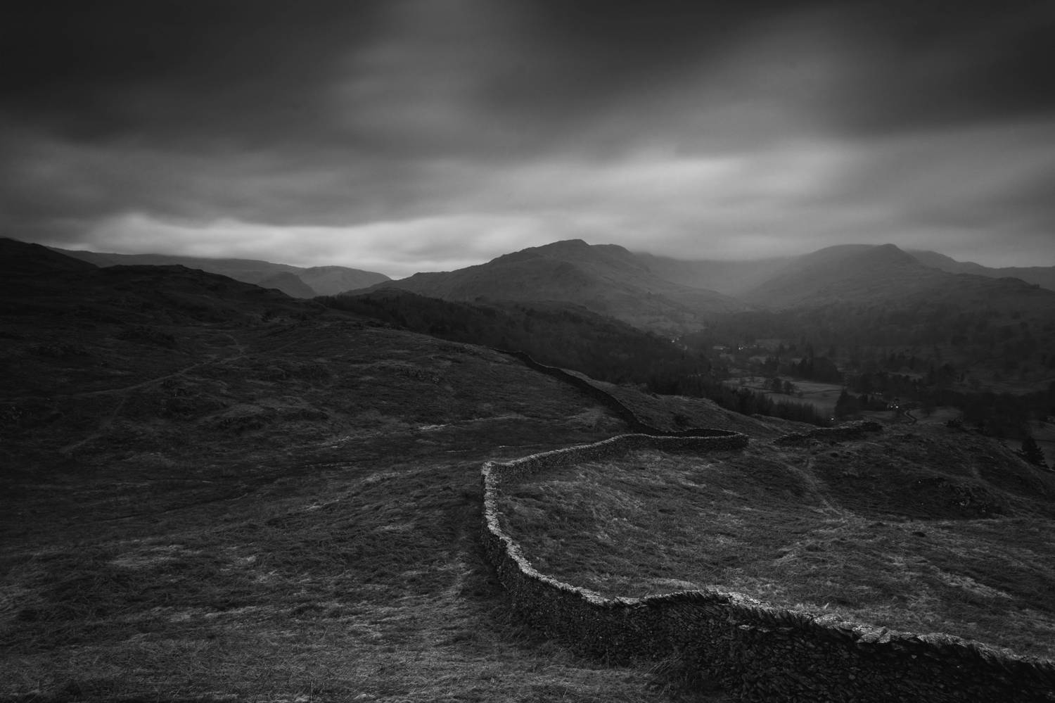 A black and white landscape photography by Joshua Wyborn depicts a grim, moody view of a low, stone farm wall in the UK.
