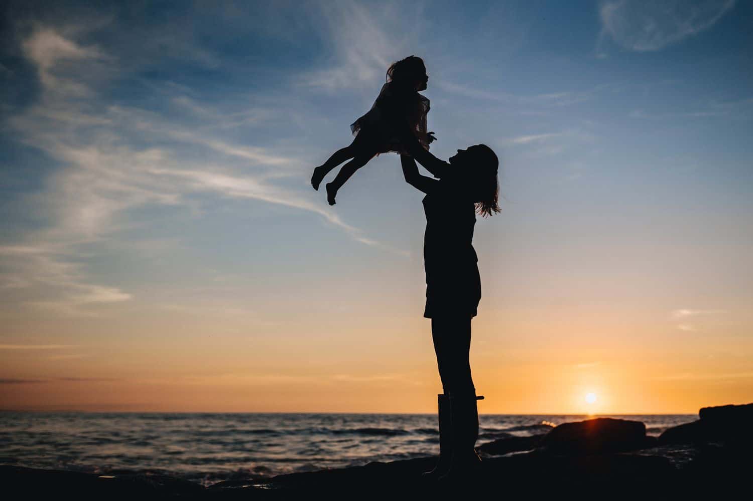 A silhouette of a woman as she lifts her child into the air at sunset on a beach.