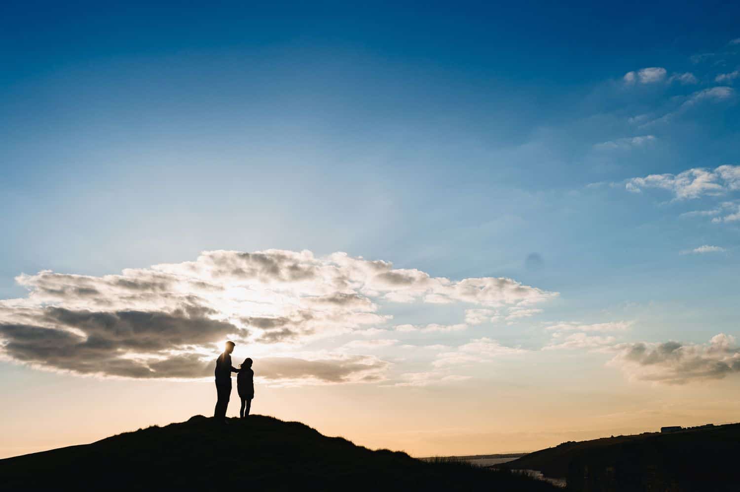 Silhouette of a father standing with his hand on his child's shoulder. They are standing on a hill overlooking the sunset sky.