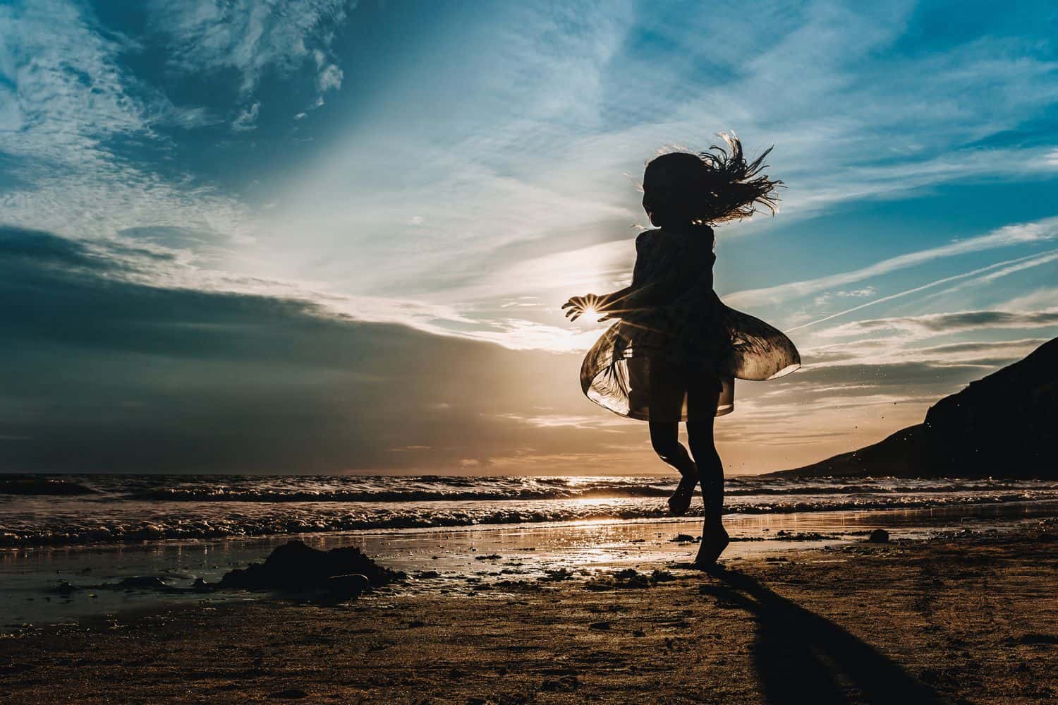 Silhouette of a little girl in a flowing dress running along the beach at sunset.