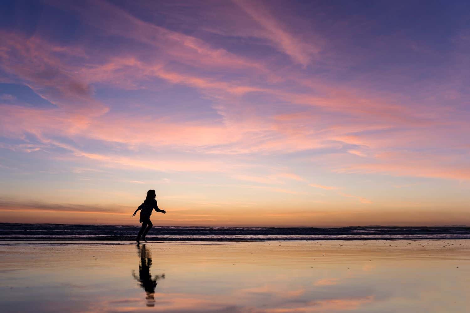 Silhouette of a child reflected in the low tide of a beach at sunset.