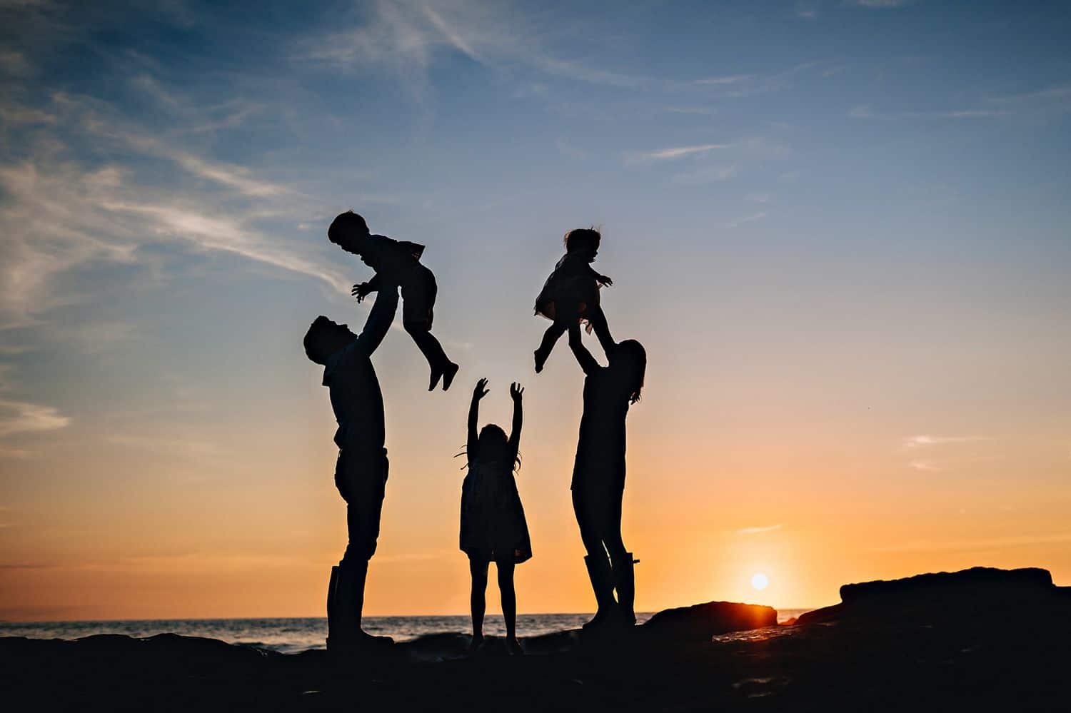 Silhouette of two parents lifting their children into the sky overlooking a beach at sunset. A third child stands in between them with her arms lifted.