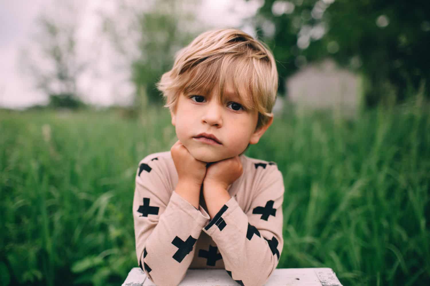A serious looking child sits in a grassy field with their chin in their hands.