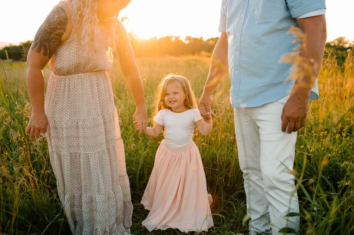 A little girl in a pink skirt holds her parents hands in a field at sunset.