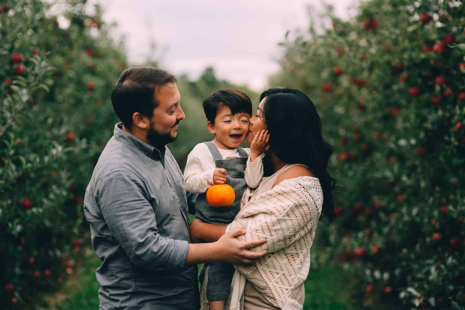 A little boy in overalls is hugged between his parents in the middle of an orchard.