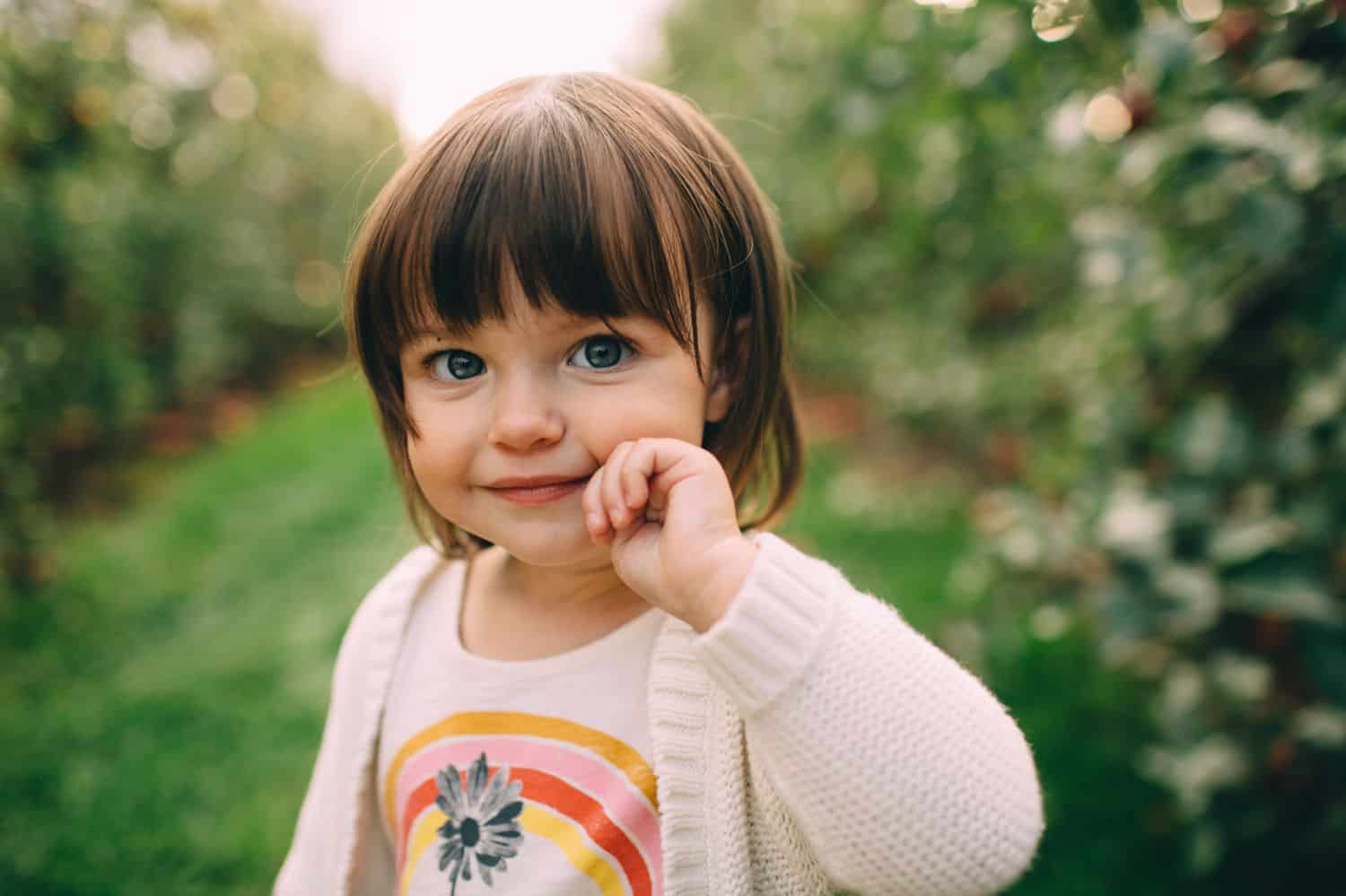A little girl wearing a rainbow t-shirt stands in an apple orchard.