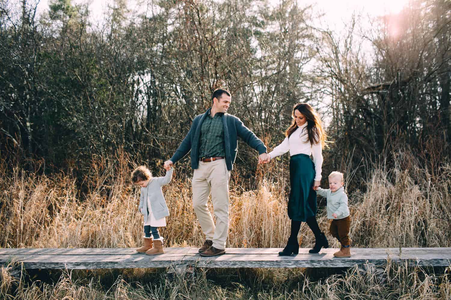 A small child leads their two parents and younger sibling along a boardwalk in a marsh surrounded by tall grass.