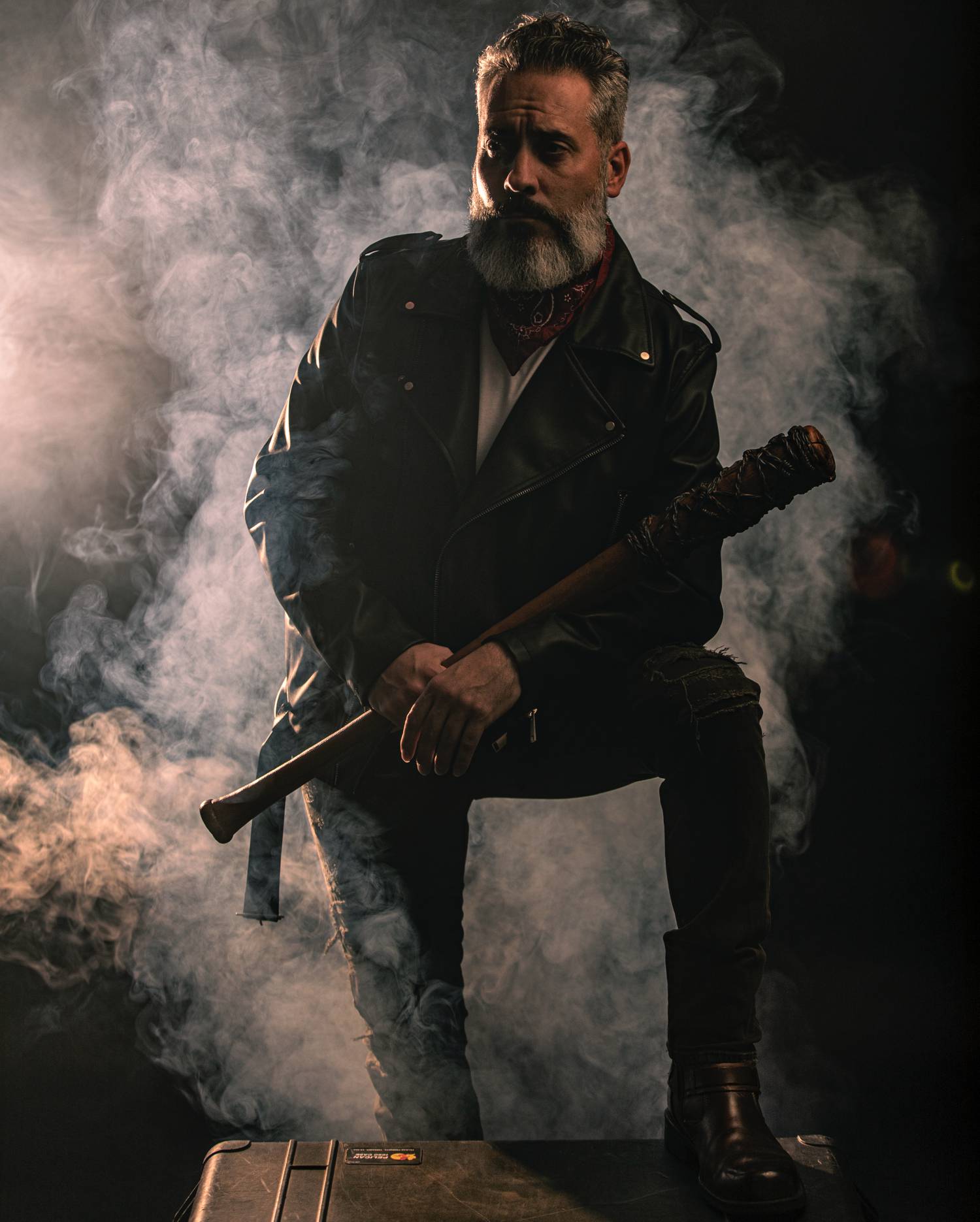 In this styled tribute photograph by Alexander Worth of Jawfox Photography, a man cosplays Negan from The Walking Dead, wearing a leather jacket and carrying the villain's barbed wire-wrapped bat, Lucille. Behind the model the air is full of dark smoke.