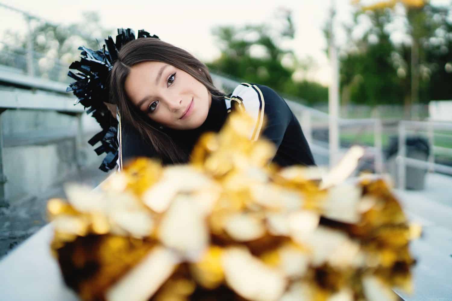 A senior girl sits beside a football field with her cheerleader pom-poms