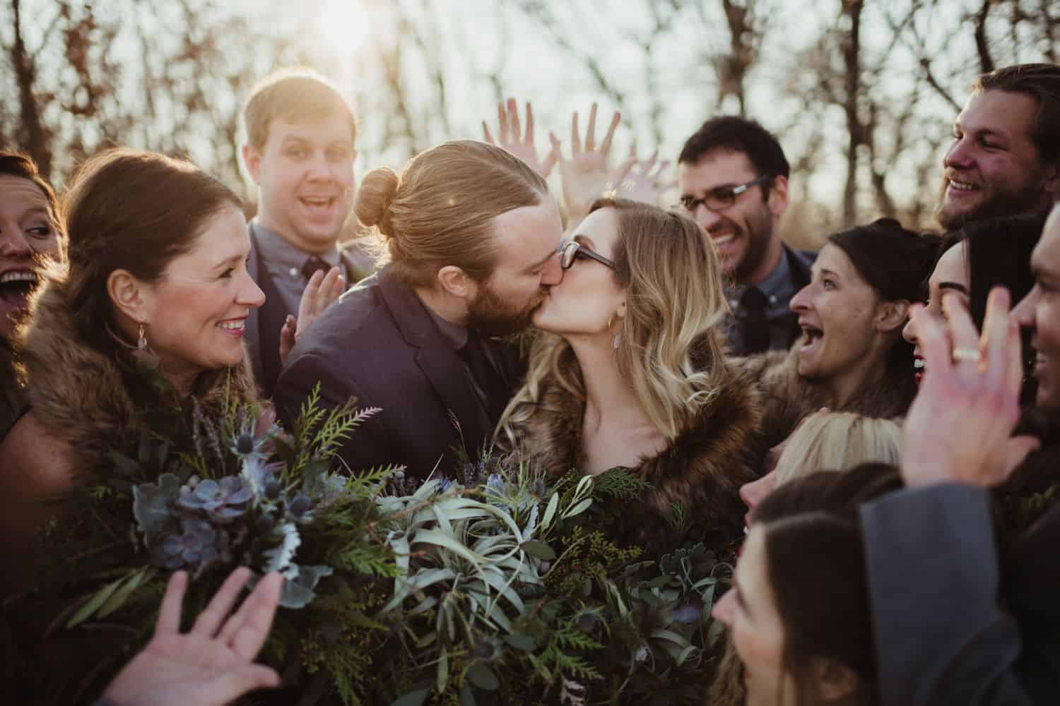 bride in a fur stole kisses her new husband while the wedding party cheers around them