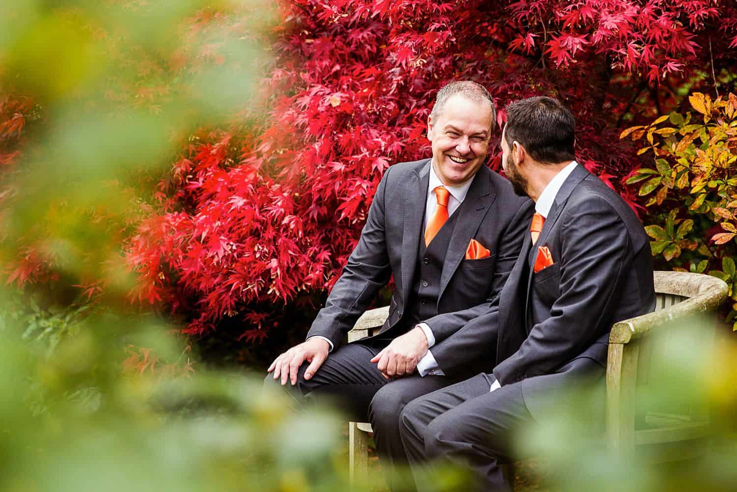 Two groomsmen sit on a bench in a church garden