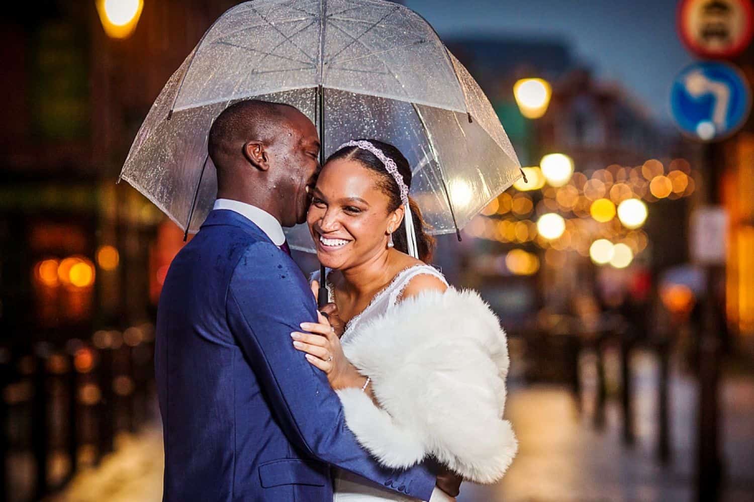 A groom in a blue tux hugs his bride in a white fur as they stand under an umbrella