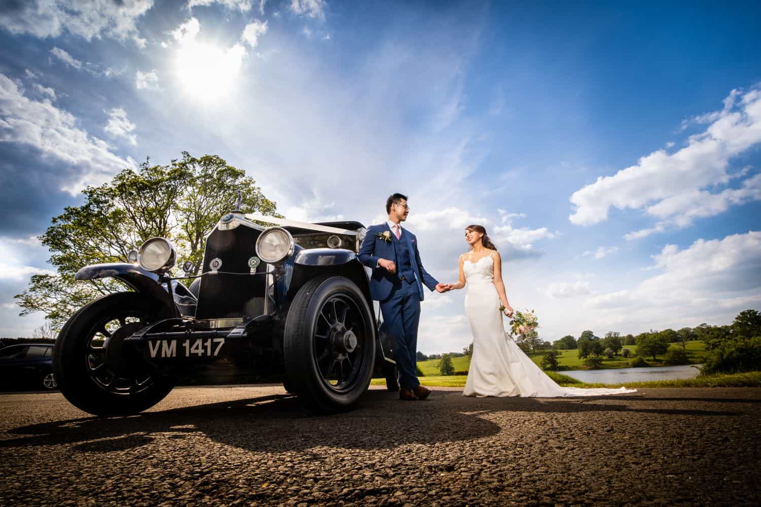 A bride and groom stand outside their getaway car on a bright sunny wedding day
