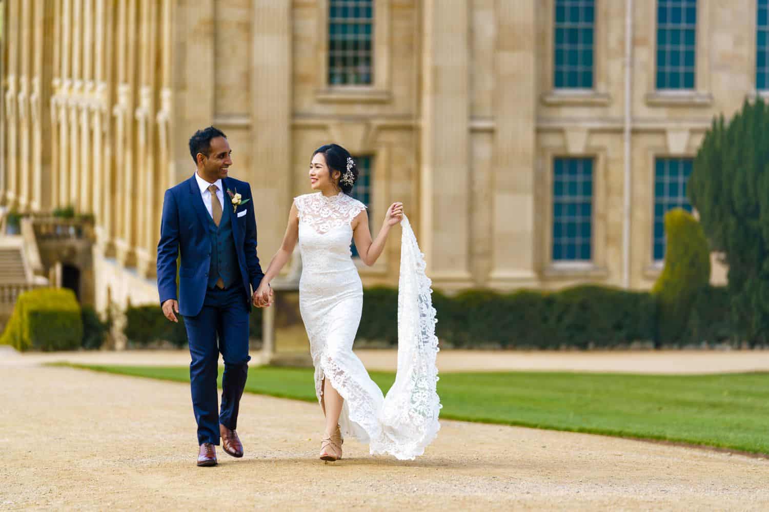 A bride and groom stroll down a garden path outside a luxurious mansion