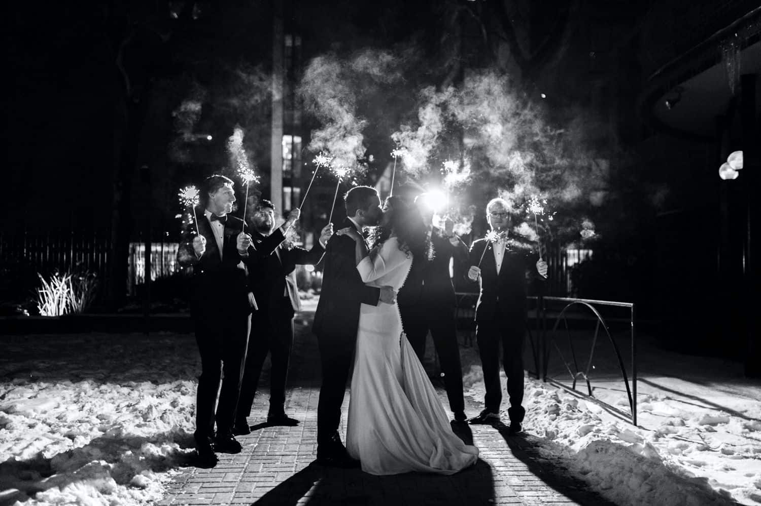 A bride and groom kiss as their wedding party waves sparklers at night