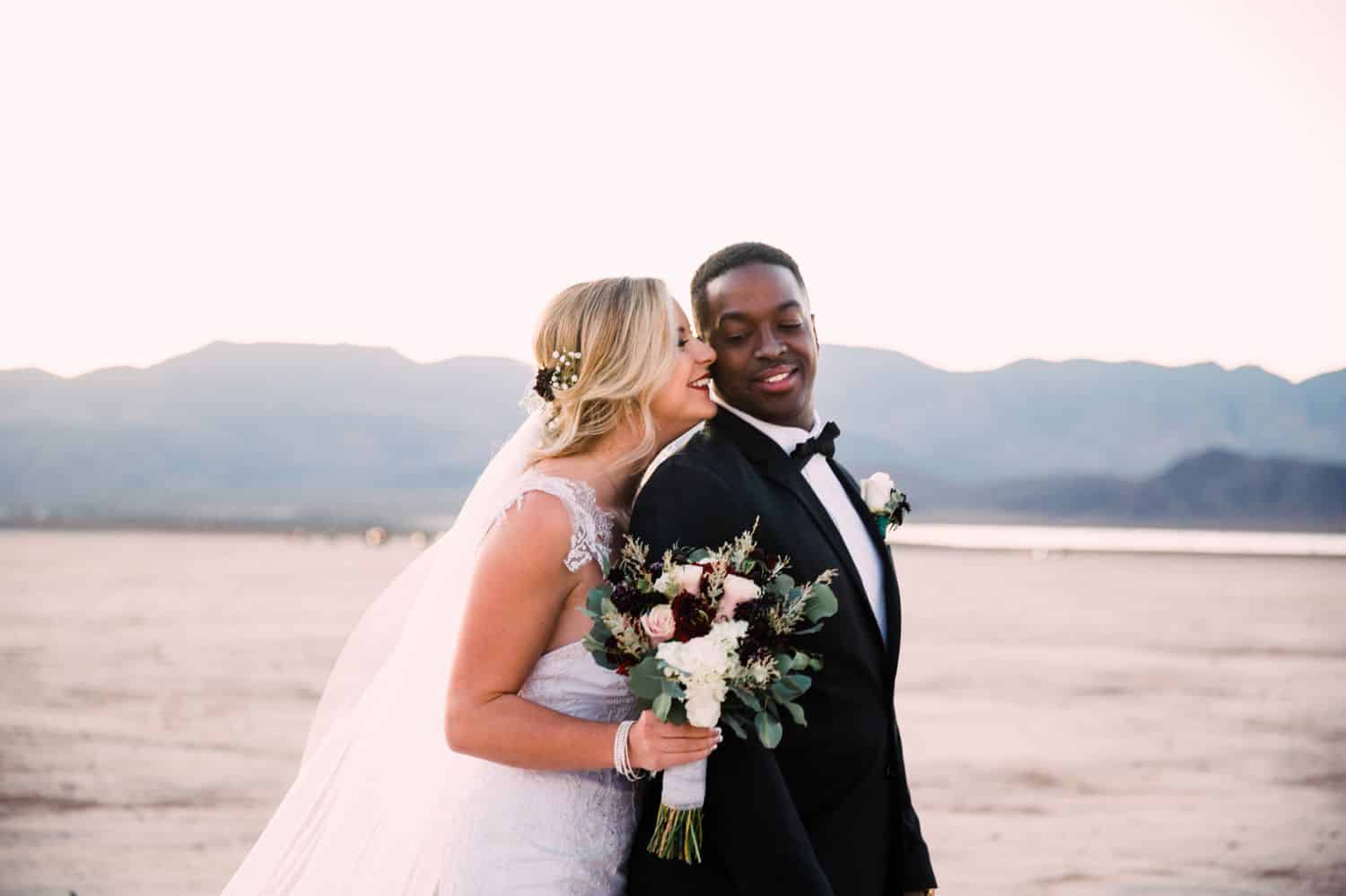 A bride stands behind the groom and kisses him on the cheek during their desert wedding
