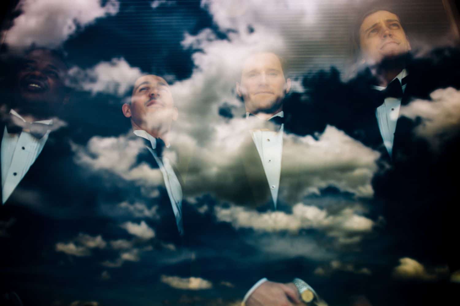 A groom and his groomsmen pose for a double exposure portrait with a deep blue cloudy sky