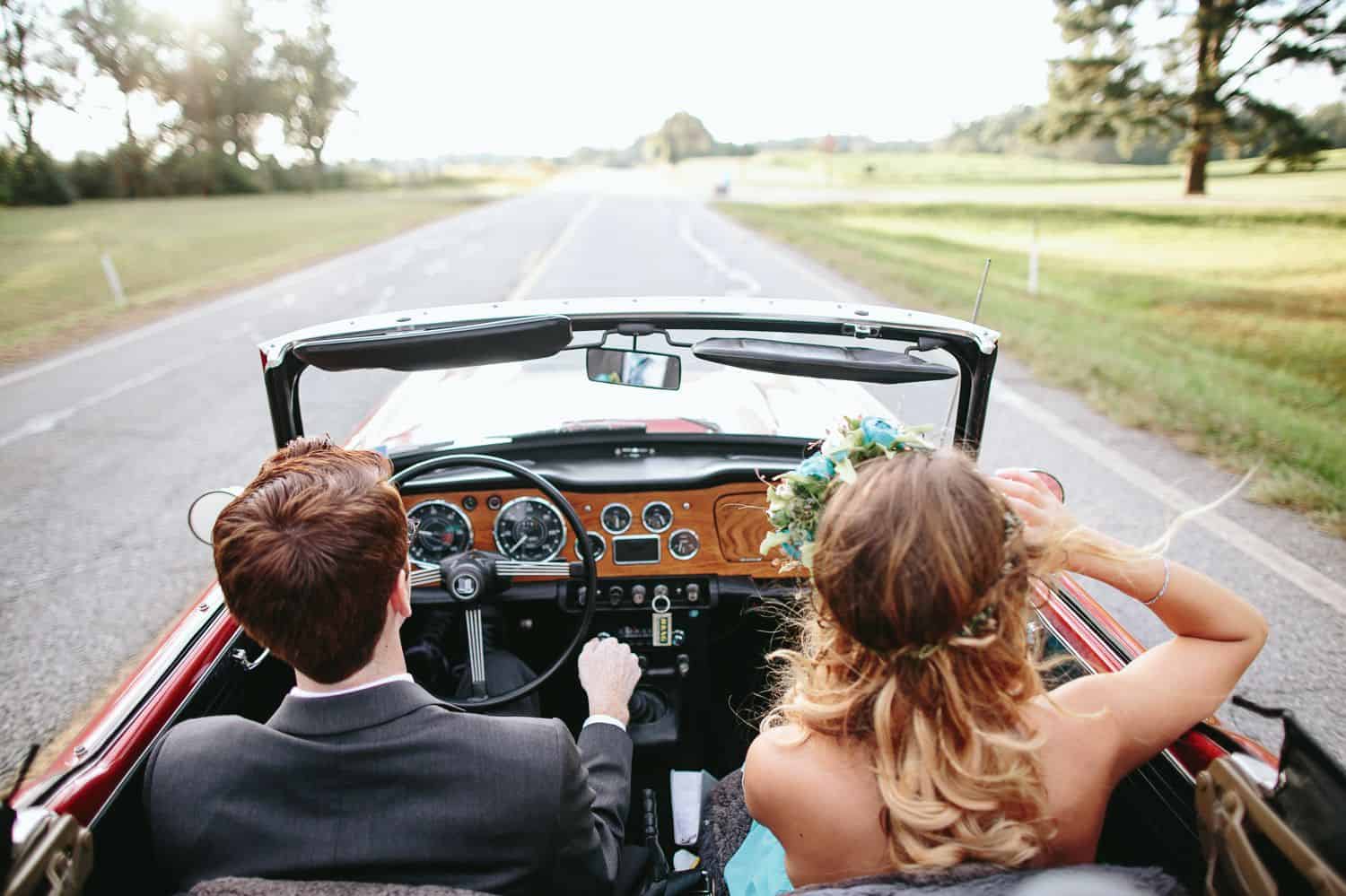 A bride and groom are photographed from behind as they drive down a country road in a vintage car