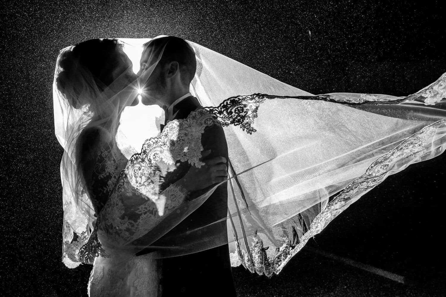 A bride's veil covers her head as well as the groom's as they pose nose-to-nose for a backlit nighttime wedding portrait