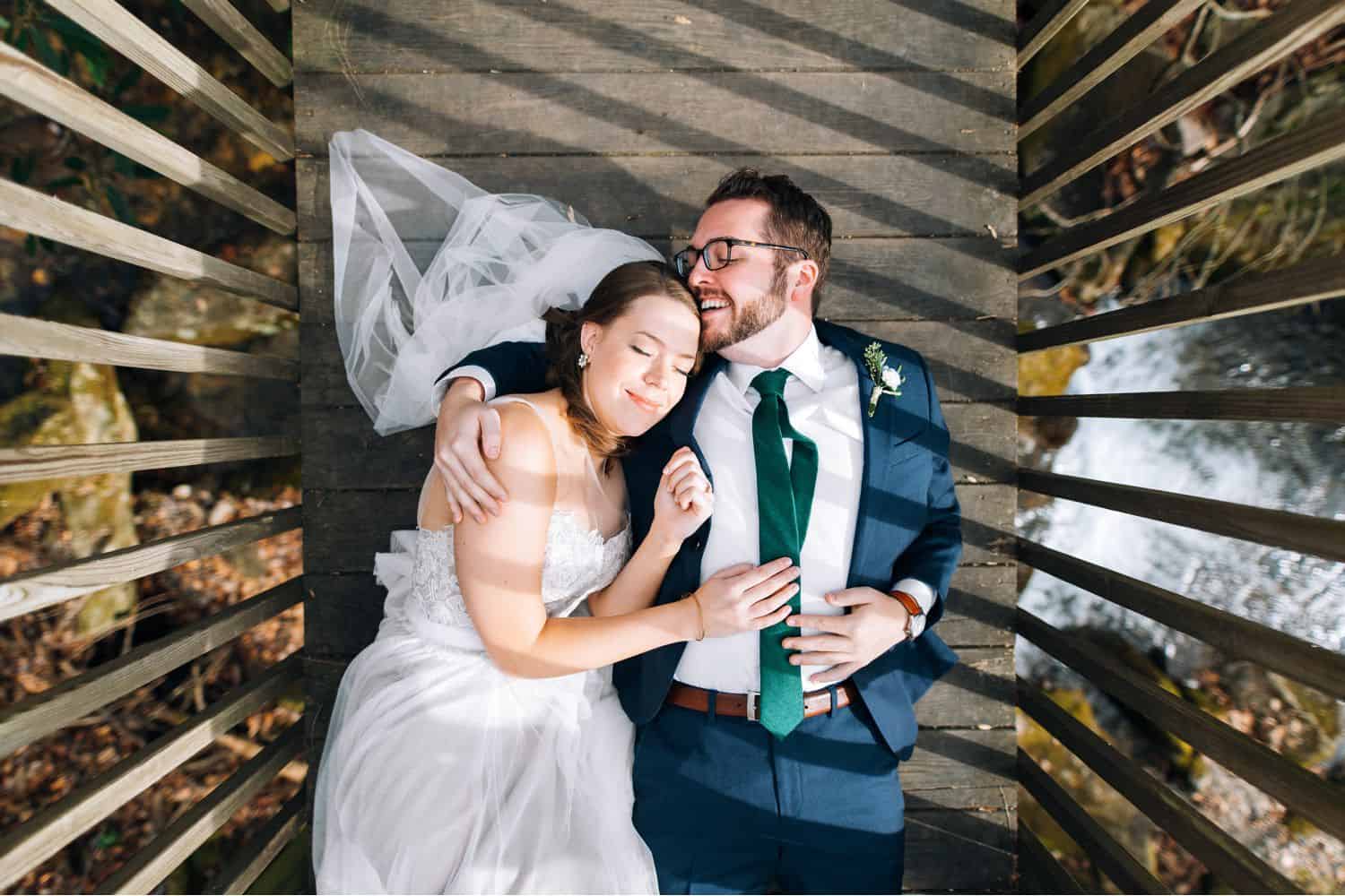 A bride and groom are photographed from above as they lie on a dock that stretches out over a lake