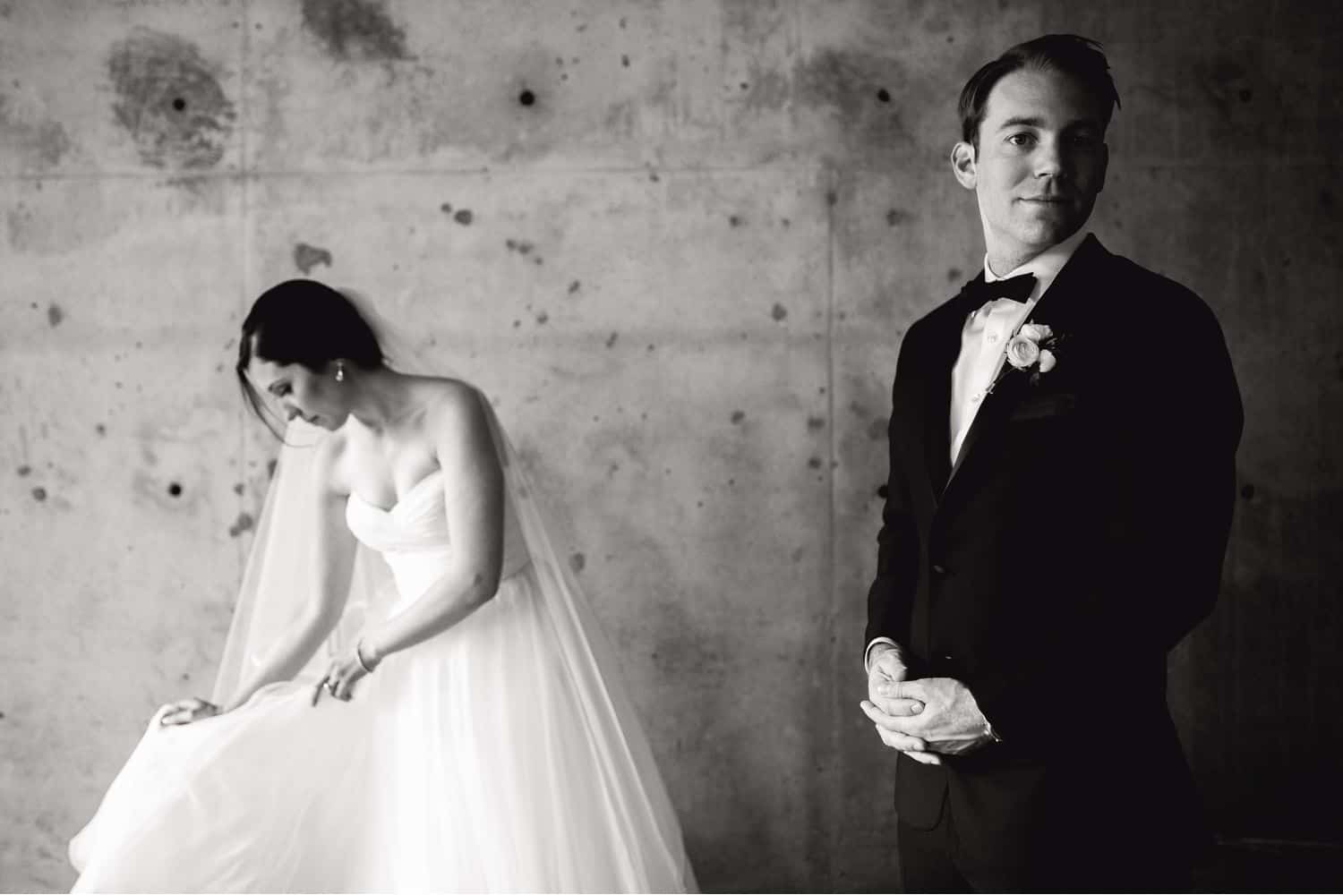 A bride adjusts her train as the groom glances at the camera in this black and white portrait from their wedding day
