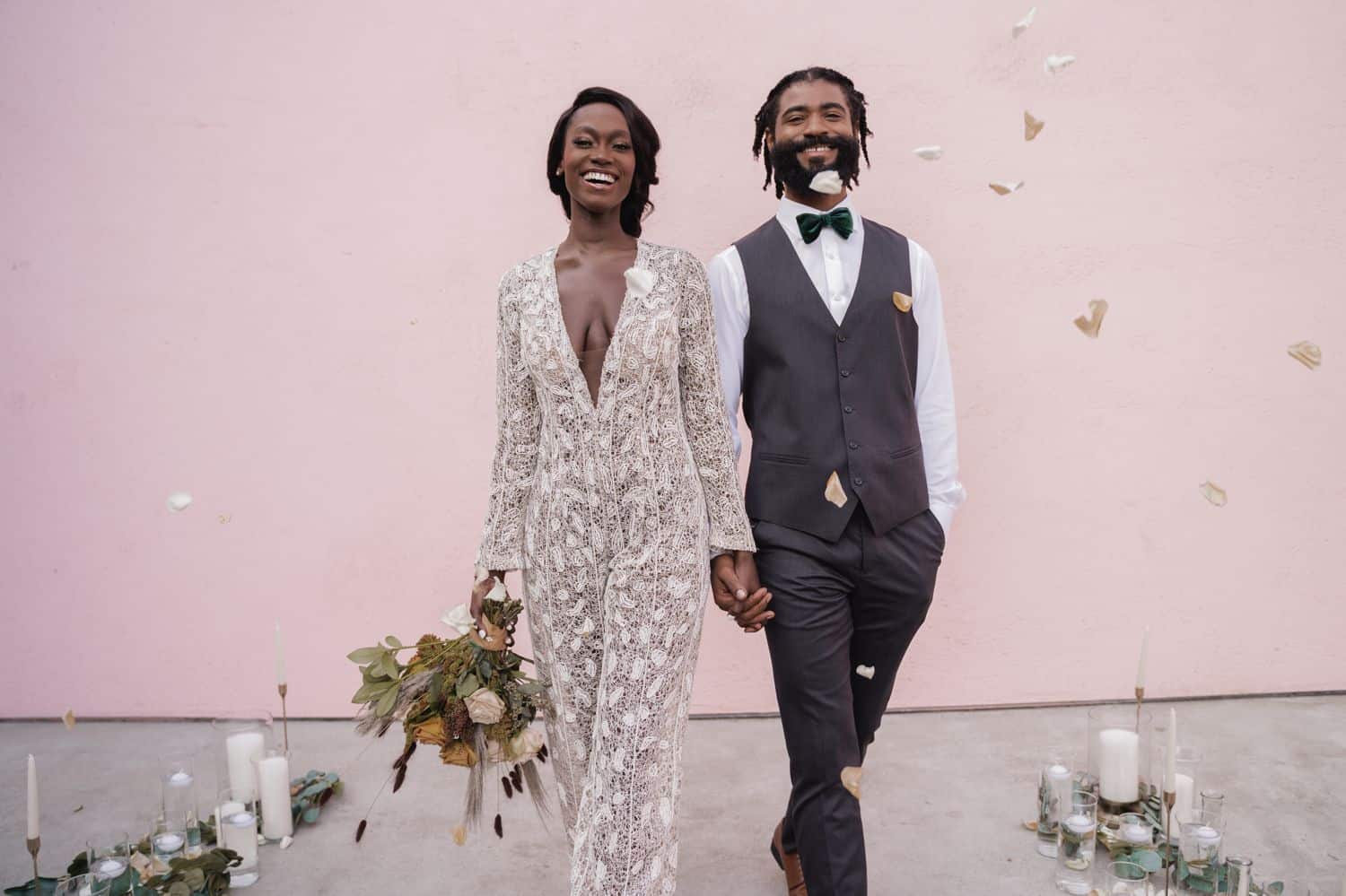 A bride in lace jumpsuit walks in front of a pink wall with her groom who is wearing a gray suit. They are in a shower of flower petals.