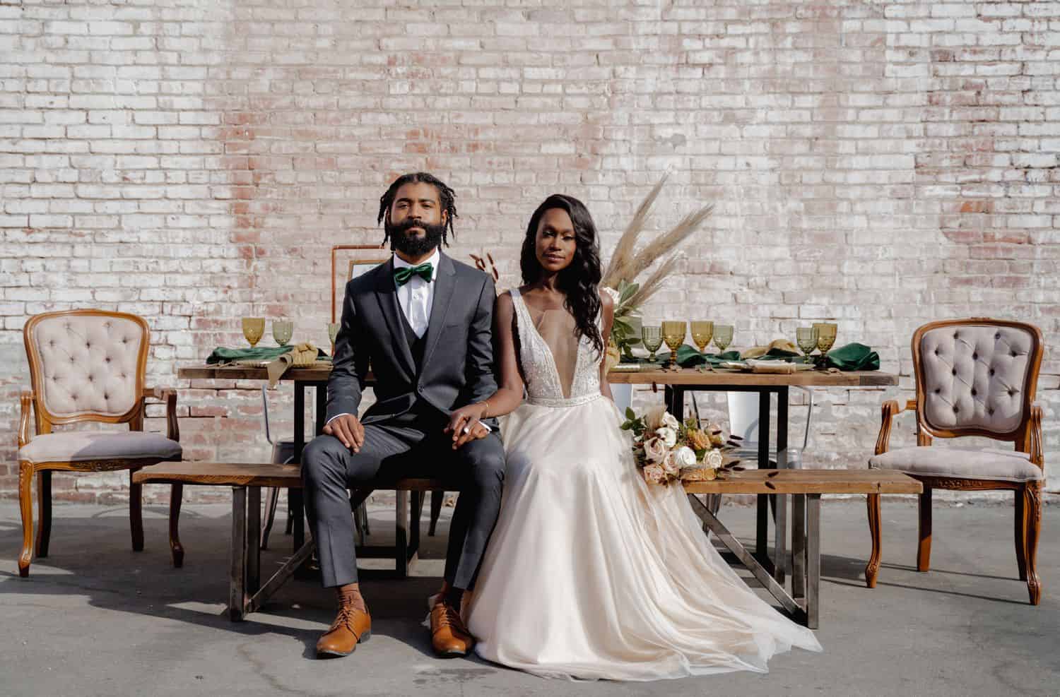A Black bride and groom sit on a decorated picnic table wearing a gray tuxedo and an ivory wedding gown in front of a brick wall.