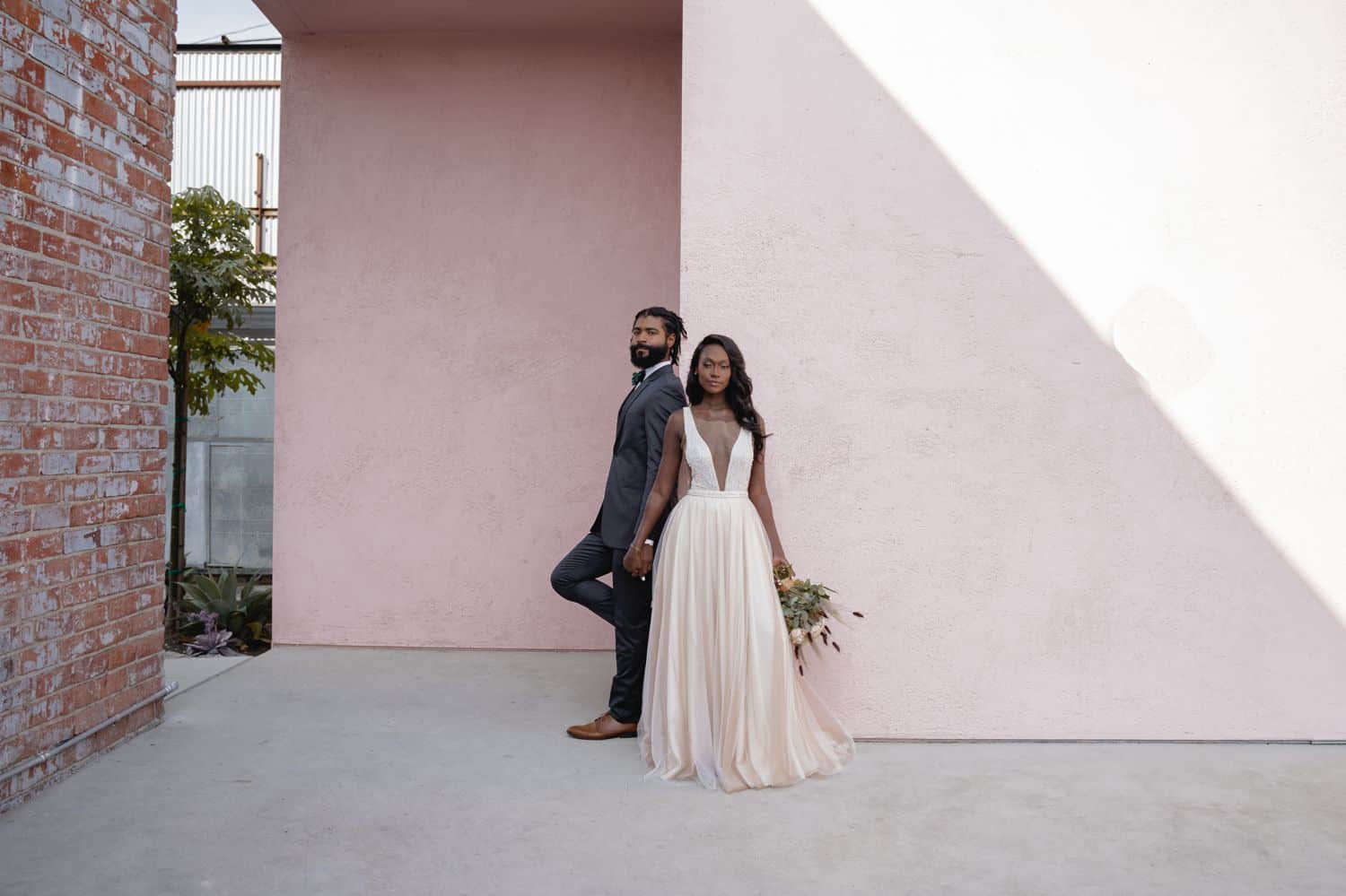 A Black bride and groom stand at the corner of a pink building beneath a slant of light.