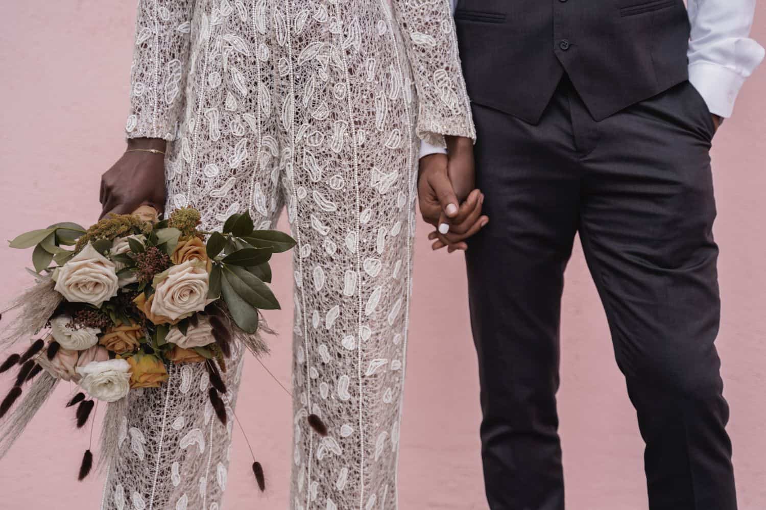 A bride in a lace jumpsuit is posed beside a groom in a gray suit. They are pictured from the waist-down in front of a pink wall.