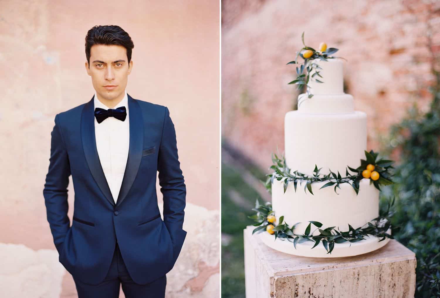 A groom in a blue tuxedo and black bow tie stands next to a tiered wedding cake, all in front of a pink wall.