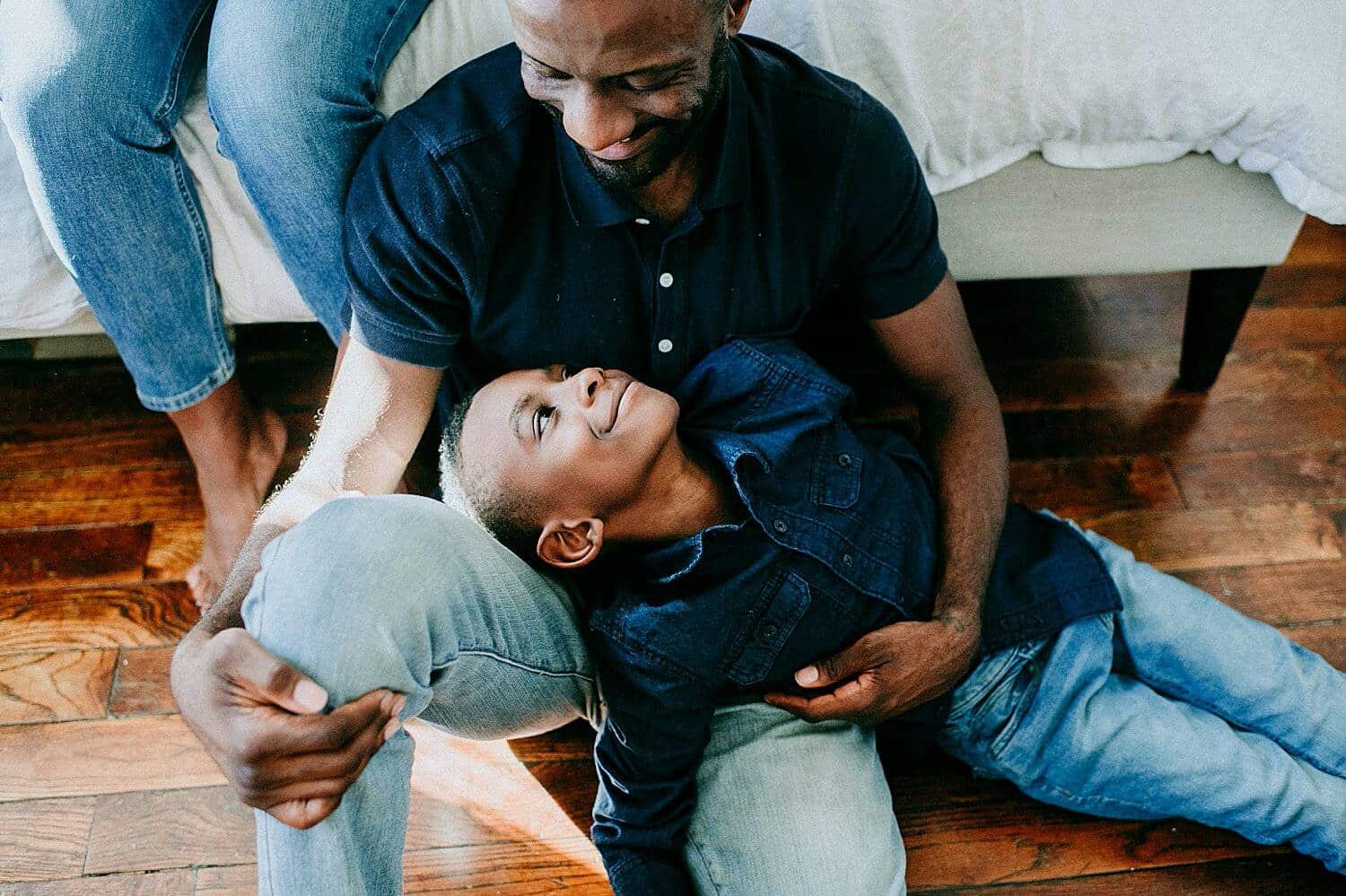Photo: Yasmina Cowan's in-home lifestyle portrait of a dad sitting on a hardwood floor and leaning against a bed as his son reclines in his lap smiling into his face.
